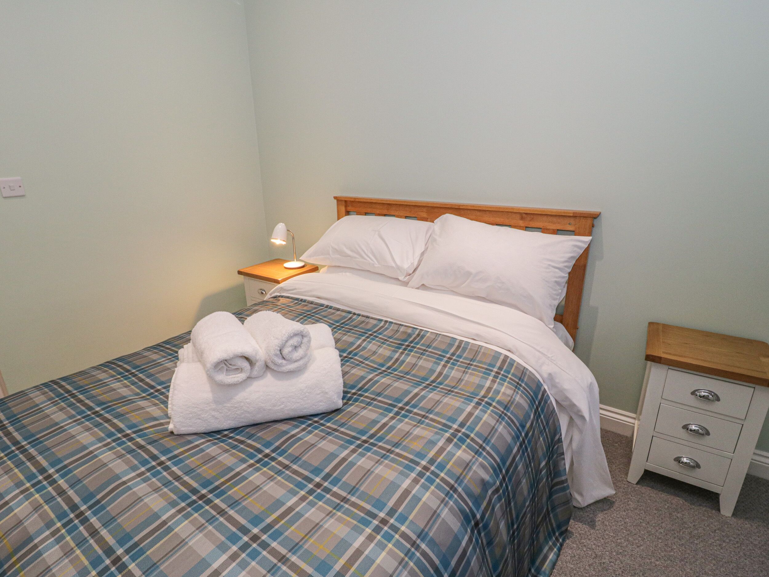A neatly made bed with a plaid bedspread, two rolled towels, a bedside lamp, and a nightstand with drawers.