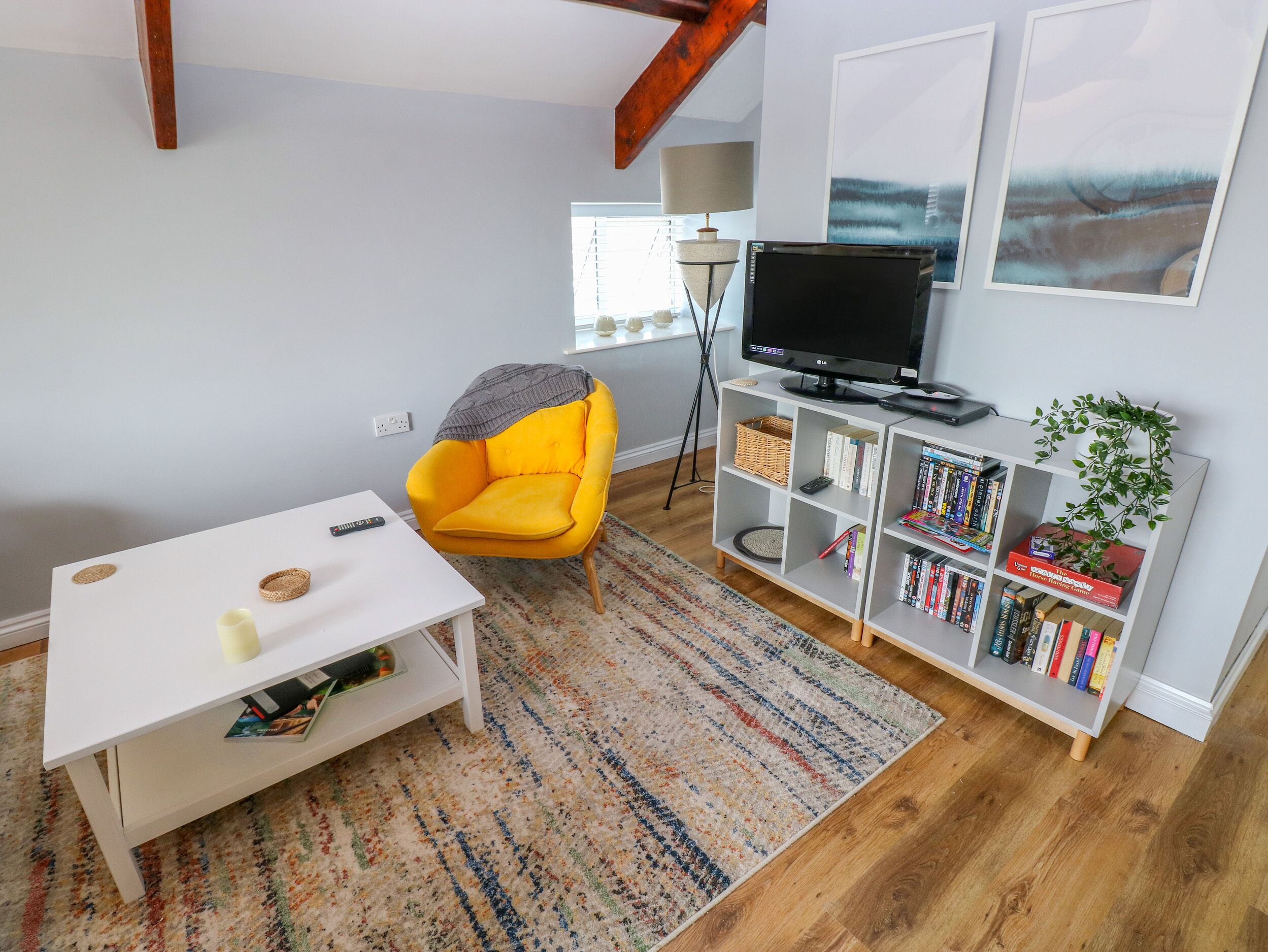 A living room with a yellow armchair, a white coffee table, a bookshelf, a television, and a window with blinds.