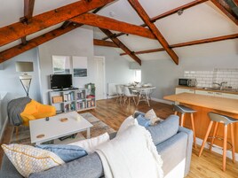 A spacious loft with a high ceiling, exposed wooden beams, a cozy living area with a sofa and coffee table, a dining area with a table and chairs, and a kitchenette with a microwave and sink.