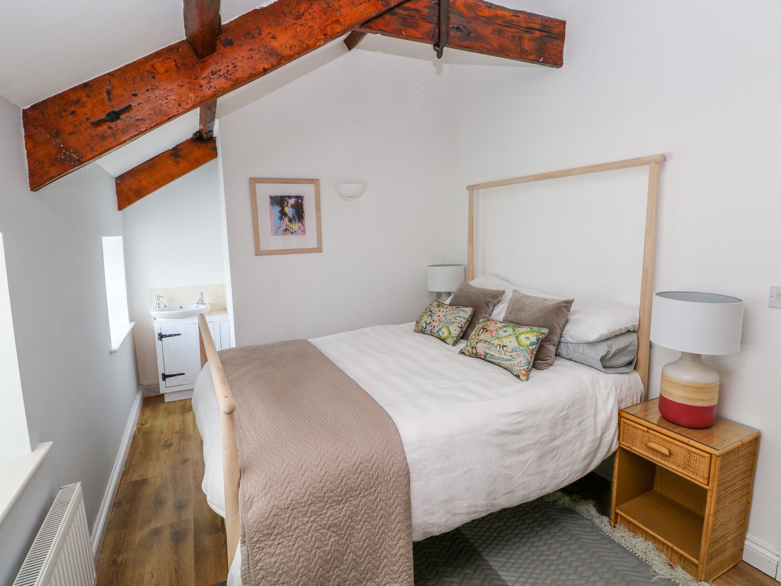 A bedroom with a bed, pillows, a bedside table with a lamp, a wooden headboard, a window, and a bathroom with a sink.