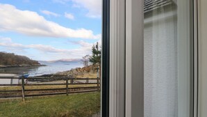 Blick aus einem Zimmer mit großem Fenster auf eine Küstenlandschaft, mit Holzzaun, Rasenfläche und einem Gewässer.