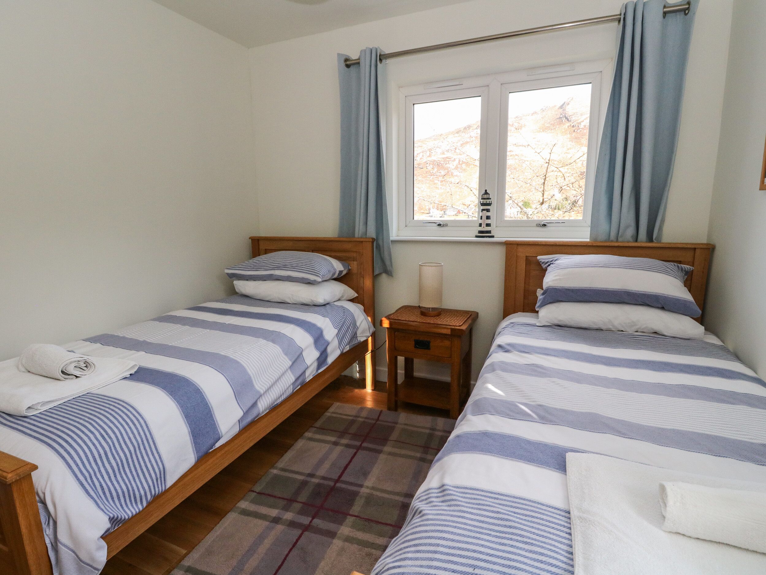 Un dormitorio con dos camas individuales, una mesita de noche con lámpara, una ventana con cortinas y una alfombra a cuadros.
