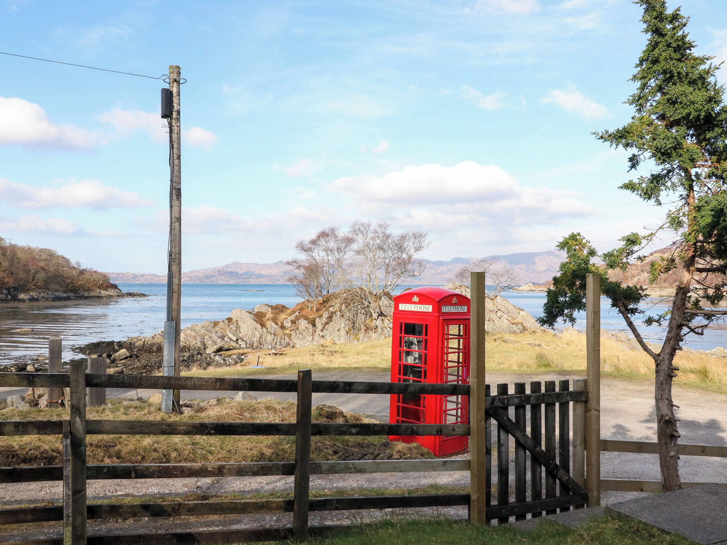 Una cabina telefónica roja junto al agua, rodeada por una valla de madera y un litoral rocoso.