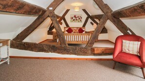 Une chambre à coucher aménagée dans un grenier, avec des poutres en bois apparentes, un lit en bois, une chaise rouge et une petite table.