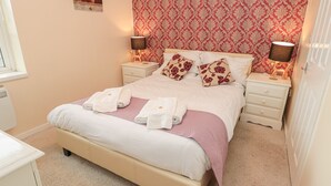 Una cama bien hecha con ropa de cama blanca y detalles en rosa, dos lámparas de noche y un papel tapiz estampado.