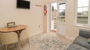 Sala de estar con un sofá gris, una mesa de madera con dos sillas, un televisor de pantalla plana en la pared y un ventanal corredero que da a un espacio exterior.