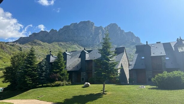 En fjällstuga med flera byggnader i chaletstil, en gräsklädd gräsmatta och en imponerande bergskuliss i bakgrunden.