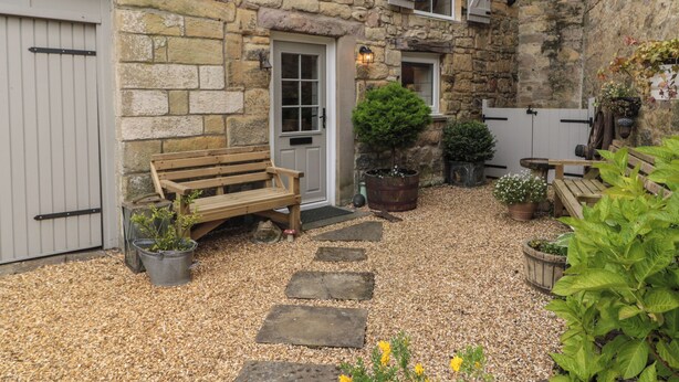 A stone building with a wooden bench, potted plants, and a gravel pathway. - The Olde Stables (Morpeth)