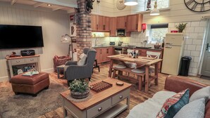 A modern kitchen with wooden cabinets, a dining area with a wooden table and chairs, a living room with a sofa and a TV, and a fireplace. - The Olde Stables (Morpeth)
