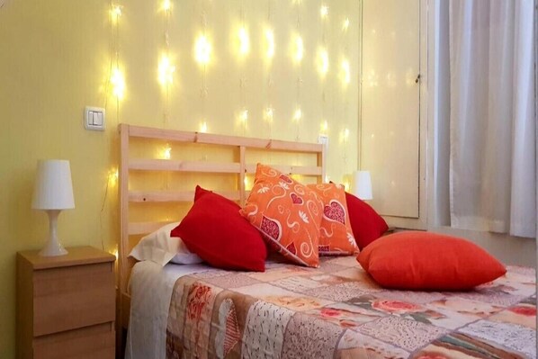 A bedroom with a wooden headboard, a bed with a patterned bedspread, and two red pillows. There is a nightstand with a lamp and a wall socket.