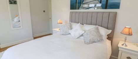 A neatly made bed with white bedding, two bedside lamps, and a painting of a pier extending into a lake.