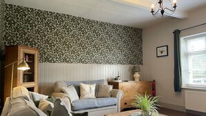 A cozy living room with a patterned wallpaper, a sofa set, a wooden cabinet, a coffee table with a plant, and a chandelier. - Warren House Cottage (Market Rasen)