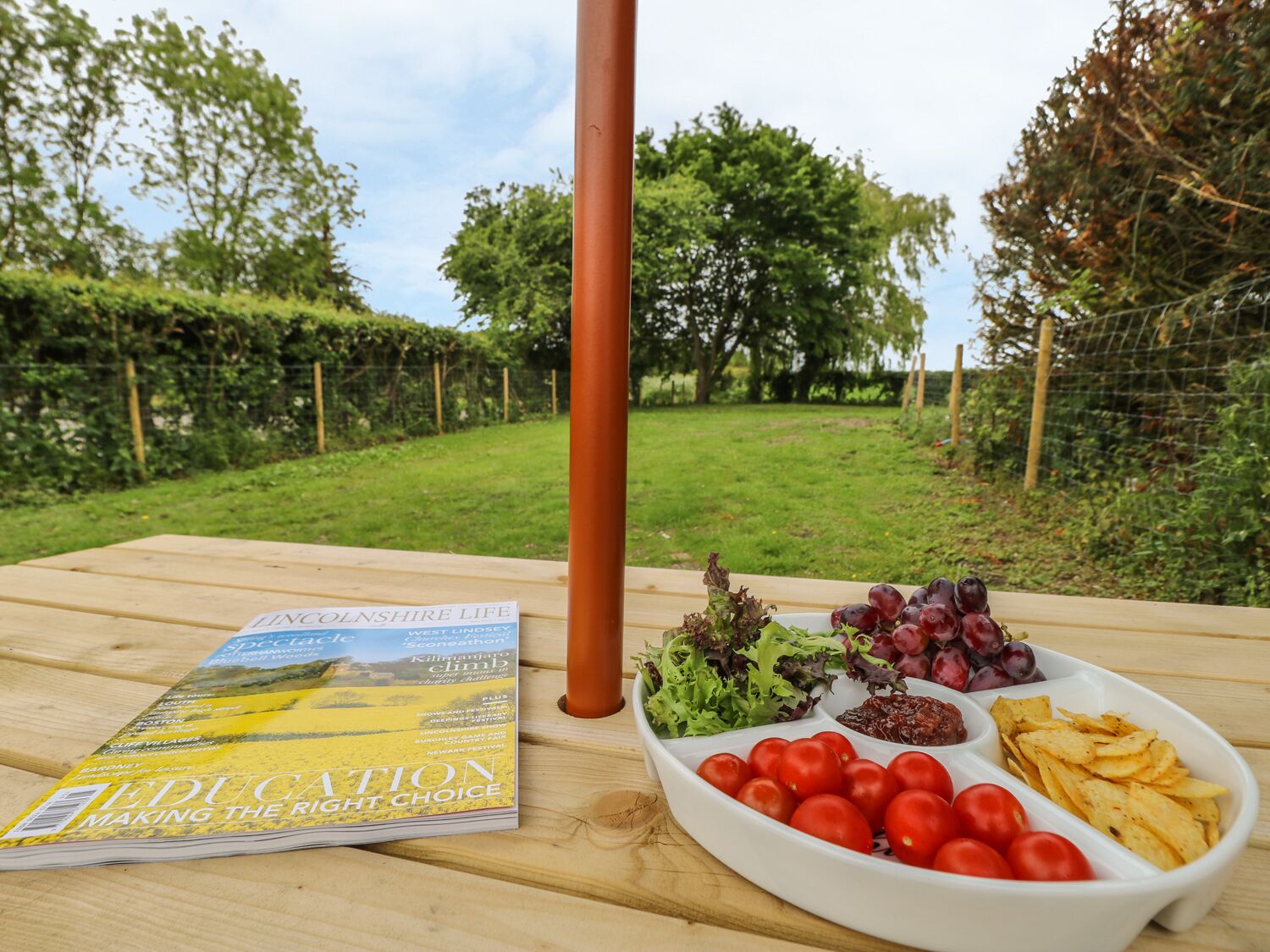 Un magazine intitulé « L’ÉDUCATION : FAITES LE BON CHOIX » est posé sur une table en bois à l’extérieur, avec vue sur un jardin.