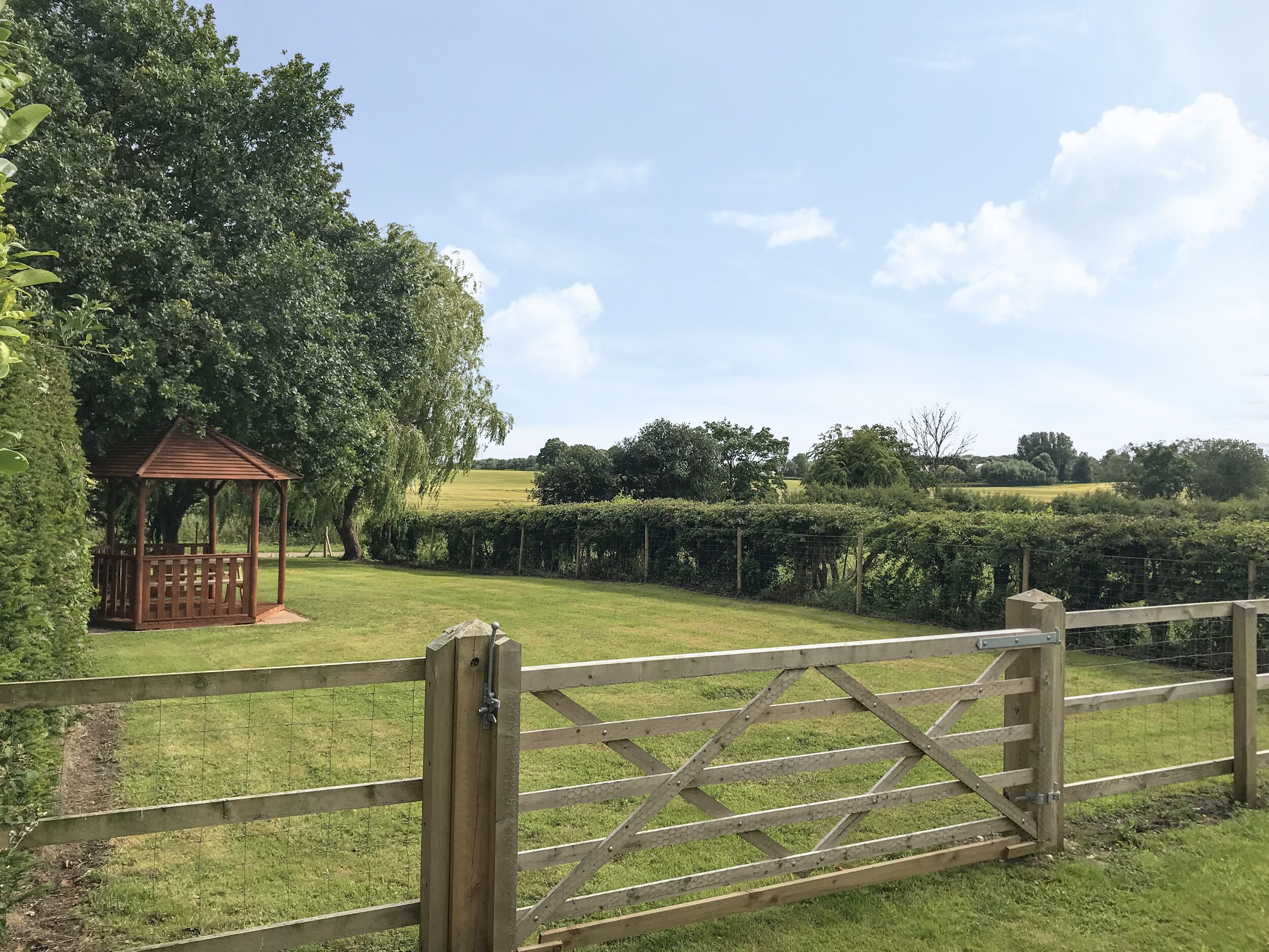 A wooden gate leading to a park with a gazebo and neatly trimmed hedges.