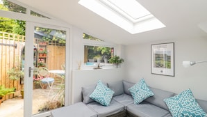 A sunlit room with a glass door leading to a garden, a grey sectional sofa with patterned cushions, and a framed picture on the wall. - Market House (Weymouth)