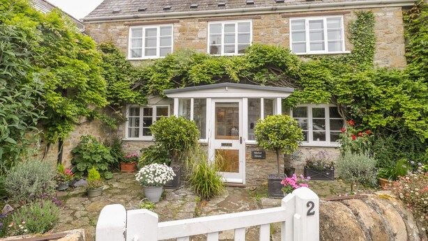 A stone house with a white door and windows, surrounded by lush greenery and potted plants. - Market House (Weymouth)