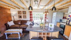 A cozy living area with a brown leather sofa, a small wooden coffee table, and a kitchen with a white refrigerator, microwave, and oven. - Gables Cottage (Market Rasen)