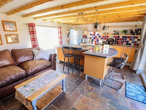 A cozy living room with a brown leather sofa, a wooden coffee table, and a kitchen area with a tiled backsplash, wooden beams, and modern appliances.