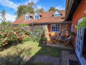 A house with a tiled roof, a brick exterior, a wooden deck with outdoor furniture, and a garden with flowering plants.