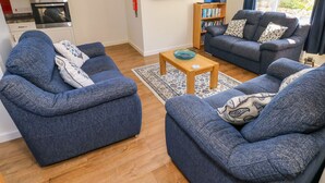 A living room with two blue sofas, a coffee table, a bookshelf, a flat-screen TV, and a kitchen area with an oven. - Little Freestone (Kilgetty)