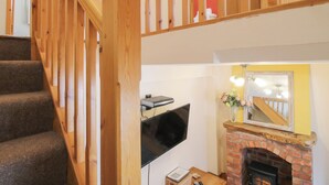 Una scala con ringhiere in legno conduce a una zona living con camino, una televisione a schermo piatto e un tavolino con un vaso di fiori.