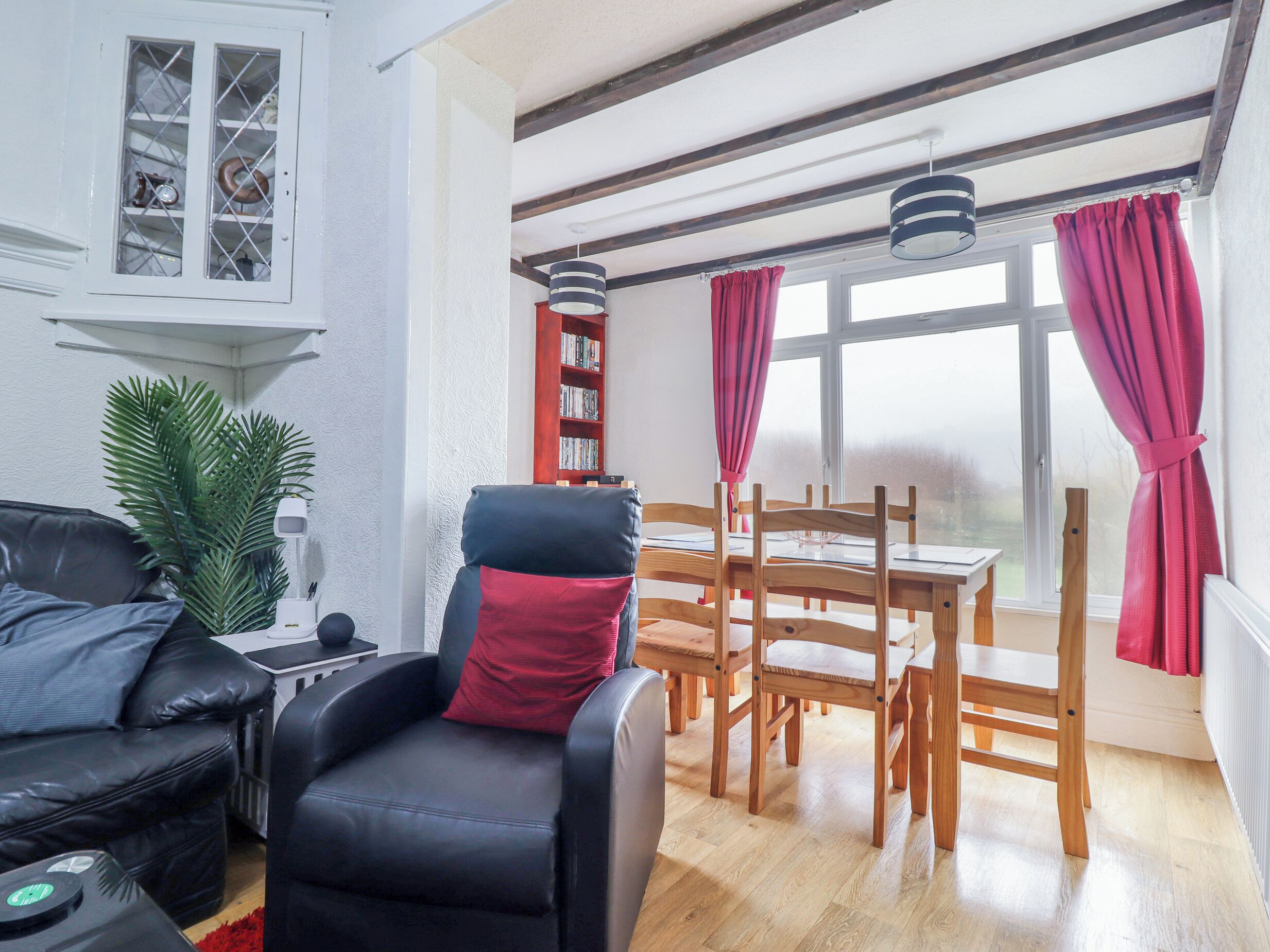 A living room with a leather sofa, a black armchair with a red cushion, a wooden dining table with chairs, a bookshelf, and a window with curtains.