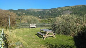 A grassy outdoor area with a wooden bench and picnic table, surrounded by greenery and hills in the background.