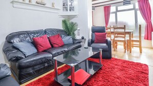 A modern living room with a black leather sofa, red and grey cushions, a glass coffee table, and a red shag rug. There is a dining area with wooden chairs and a table, and a window with red curtains.