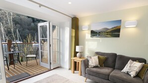 Ein Wohnzimmer mit grauem Sofa, einem kleinen Tisch und Blick auf einen Balkon mit Tisch und Stühlen.