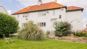 Une maison blanche à deux étages, avec un toit recouvert de tuiles rouges, un jardin avec une pelouse et une petite table entourée de chaises, ainsi qu’un mur en pierre.