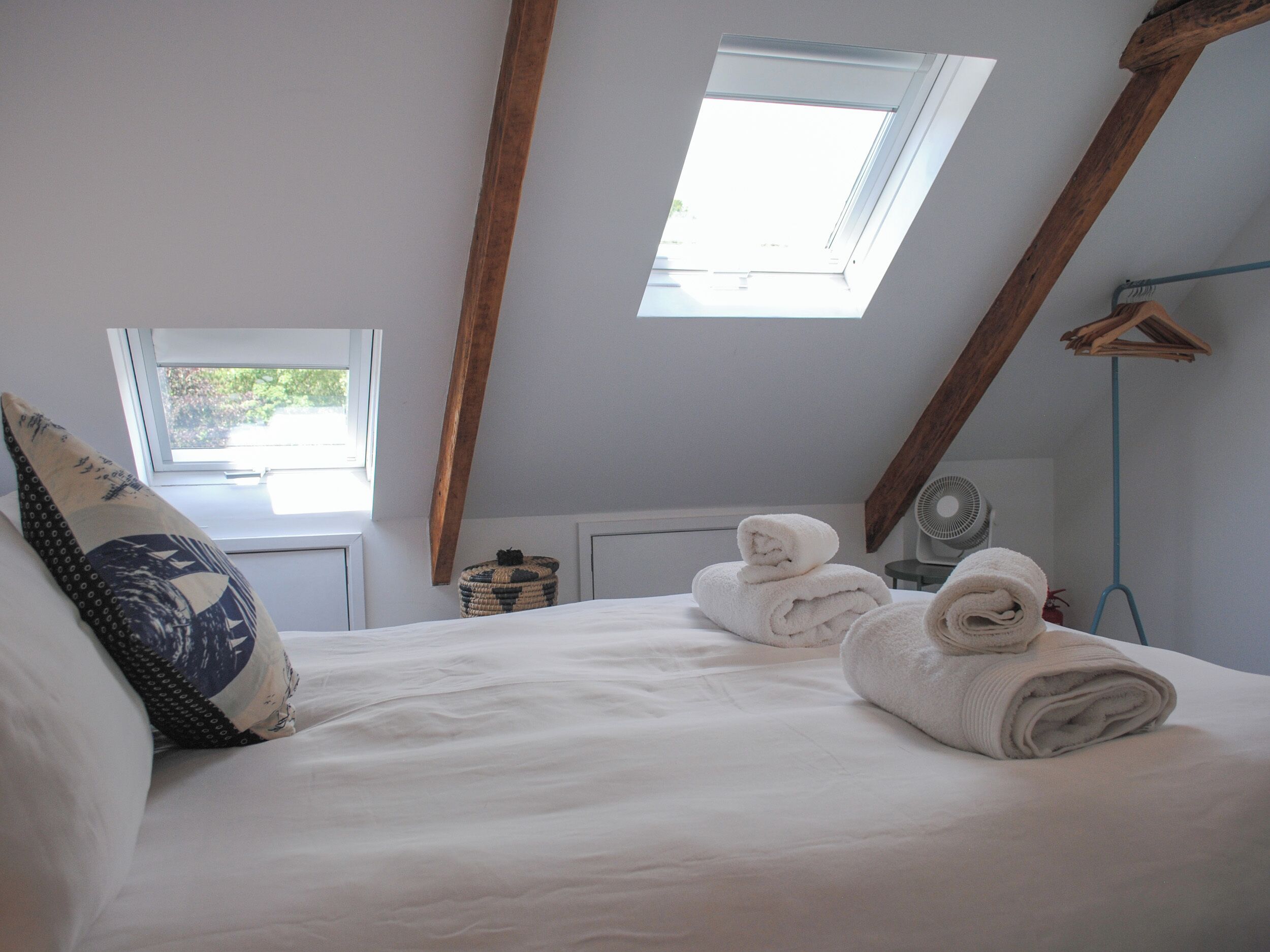 Un dormitorio con techo inclinado, una cama con ropa blanca, una ventana y un ventilador.