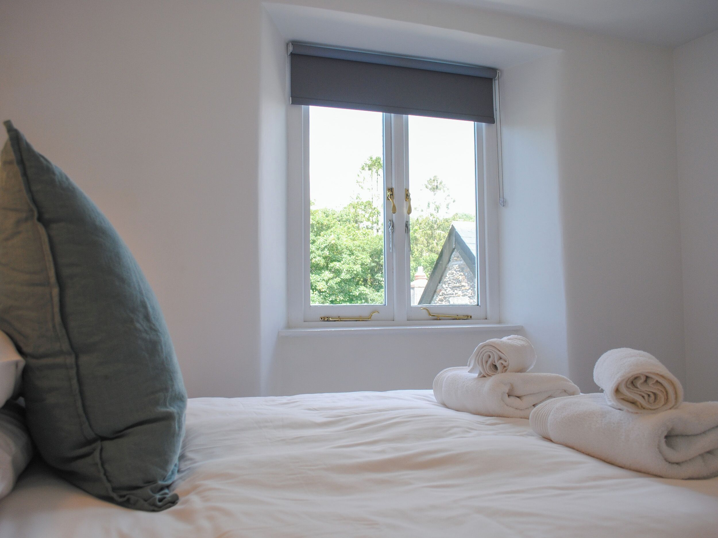 A bedroom with a bed, a window with a view of greenery, and two rolled towels on the bed.