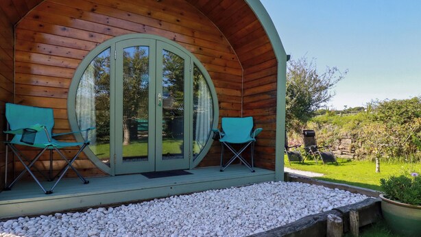 A wooden cabin with a circular door and two folding chairs on a gravel platform. - The Lamb Shack (Bodmin)