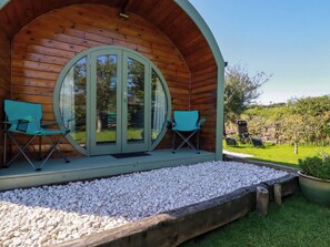 A wooden cabin with a circular door and two folding chairs on a gravel platform. - The Lamb Shack (Bodmin)