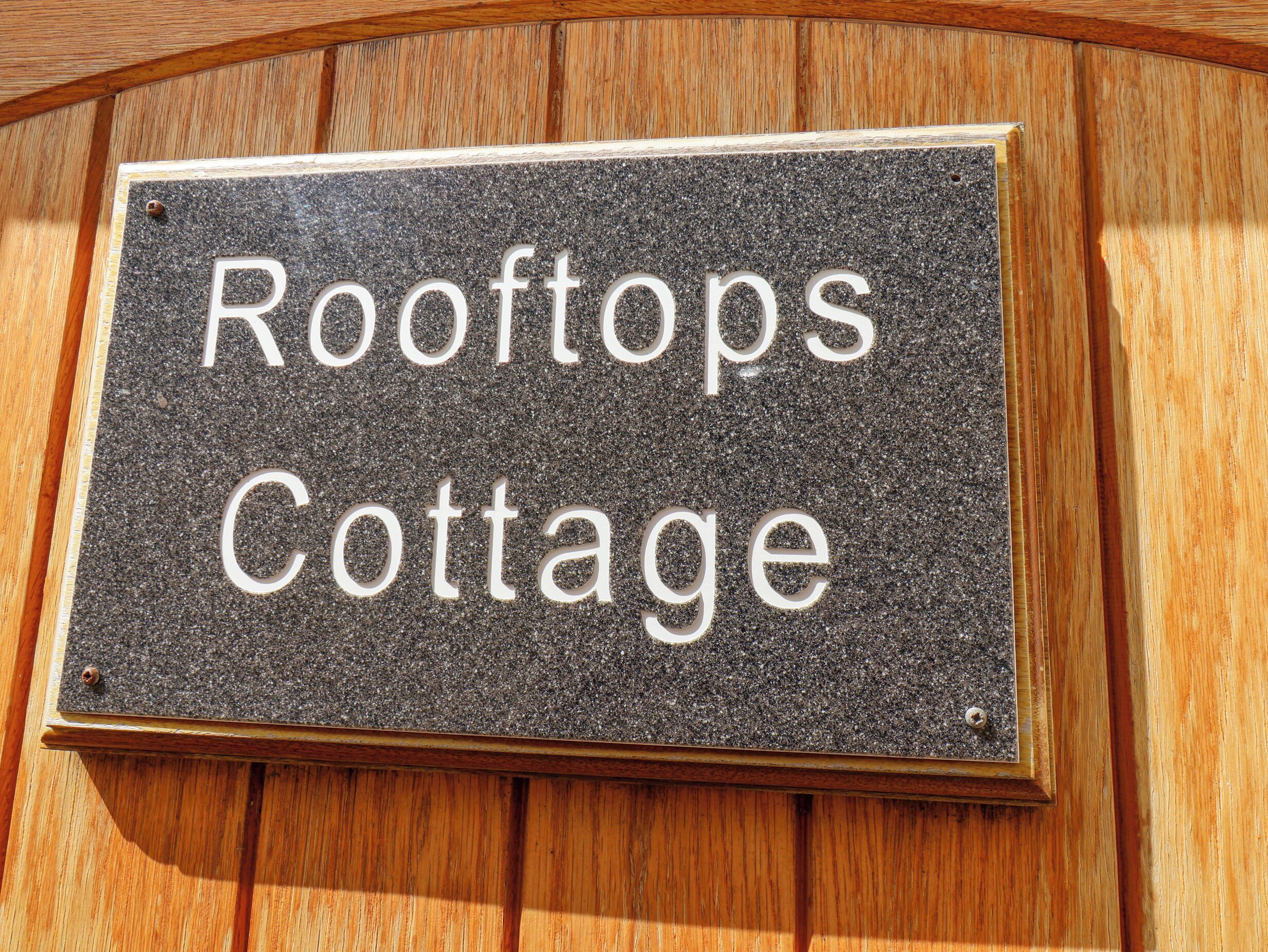 A sign with the text "Rooftops Cottage" on a textured background, mounted on a wooden surface.