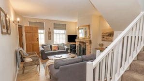 Ein geräumiges Wohnzimmer mit grauem Sofa, einem Holztisch und einem Kamin. Eine Treppe mit weißer Geländer führt zu einer oberen Ebene.