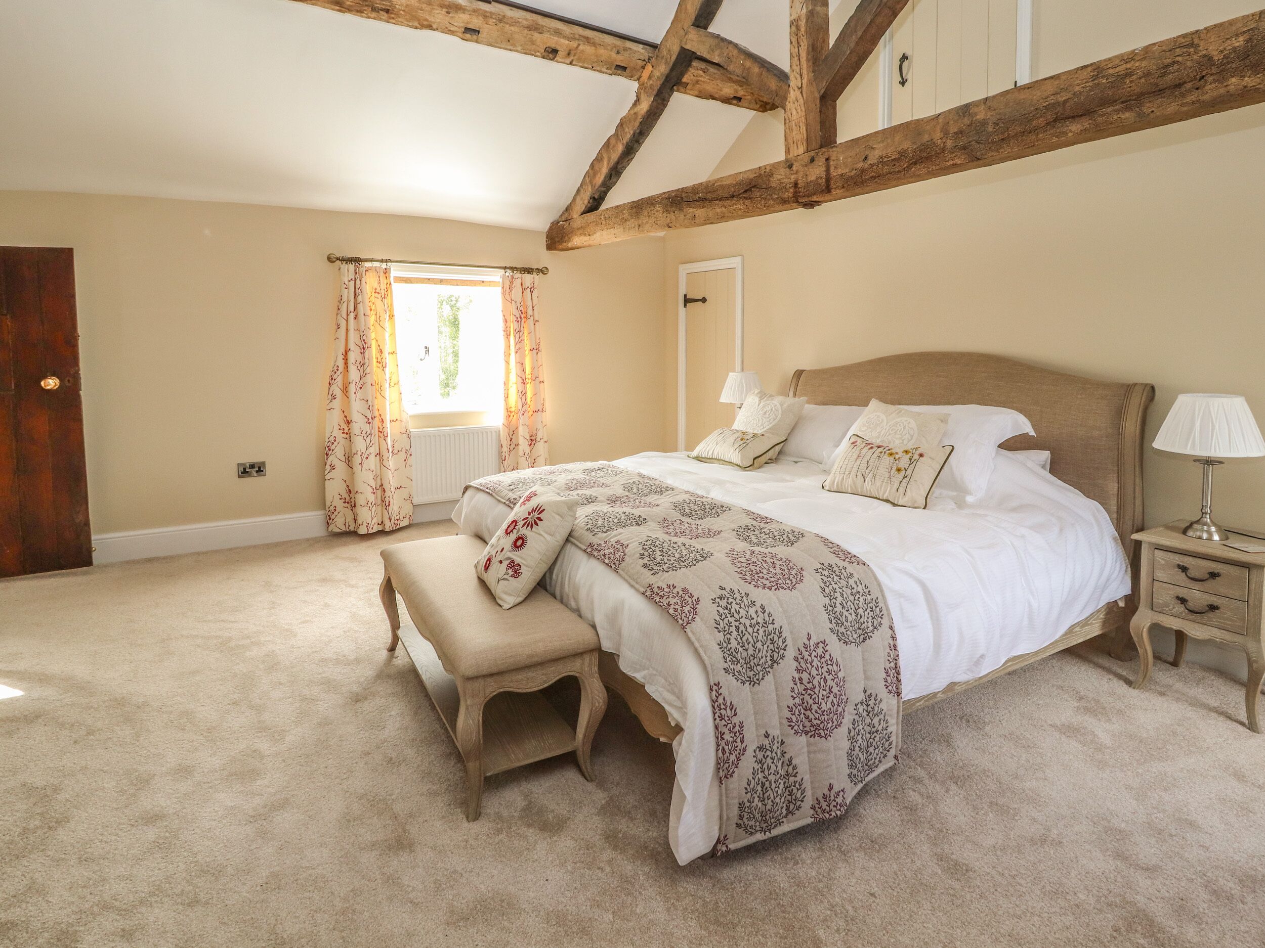 A bedroom with a large bed, a bench, a nightstand, and a window with curtains.
