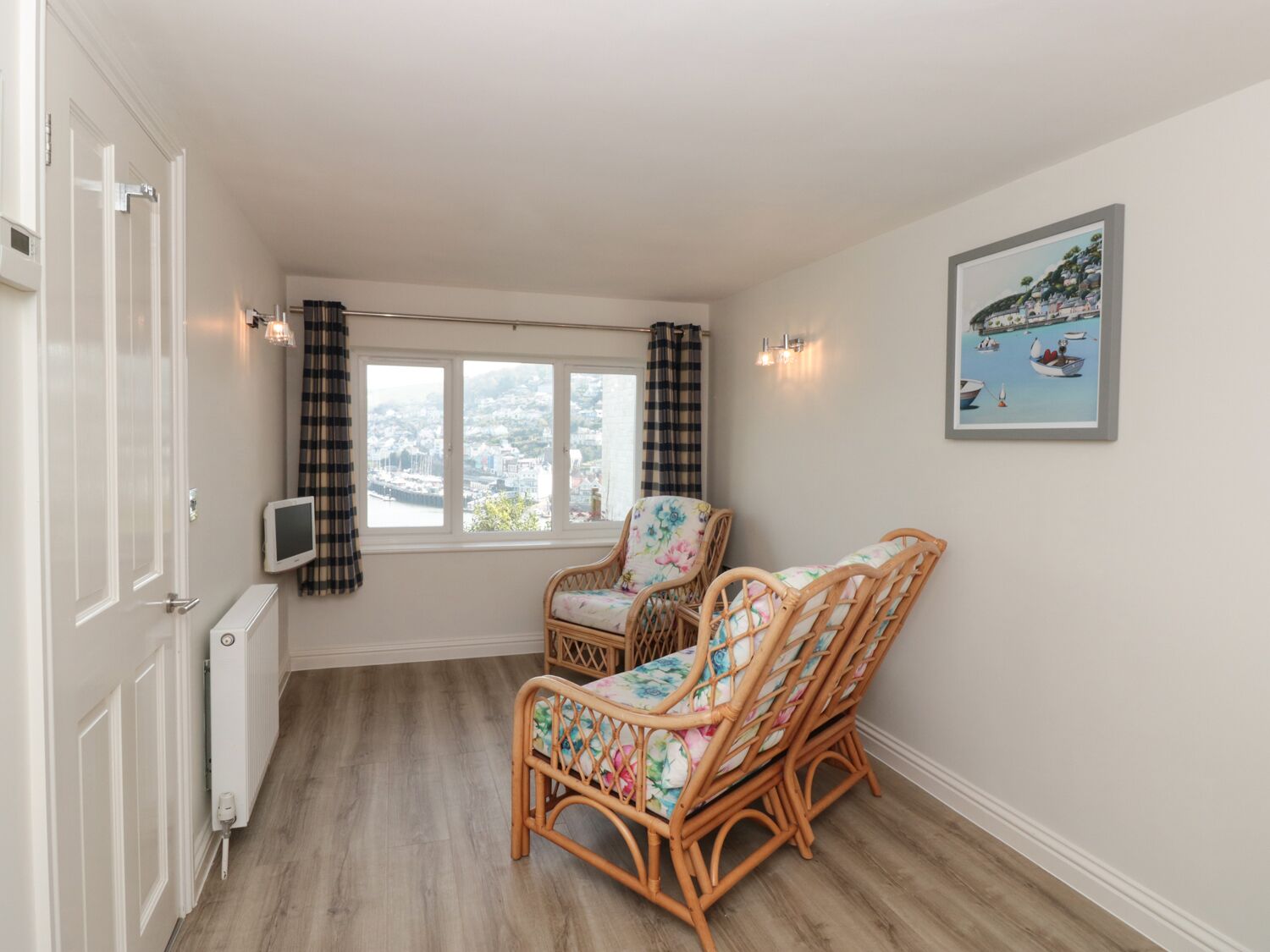 Una habitación con vista al puerto, dos sillones de mimbre con cojines estampados, un radiador blanco y un cuadro en la pared.