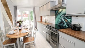 A modern kitchen with a dining area, featuring a wooden table, white chairs, stainless steel appliances, and a view of the outdoors.
