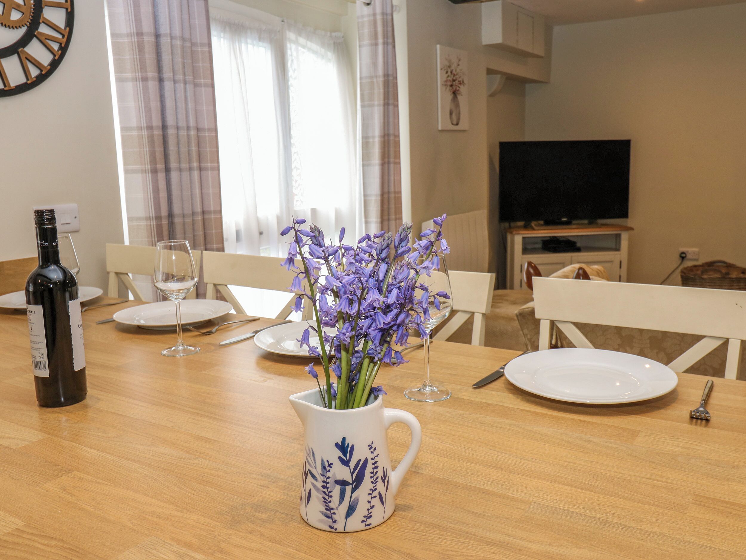 Un coin repas avec une table en bois, des assiettes blanches, des verres à vin, une bouteille de vin et un vase rempli de fleurs violettes.