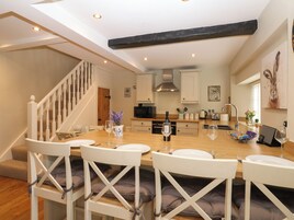 Una cocina moderna con armarios blancos, encimera de madera y horno empotrado. Hay una zona de comedor con sillas blancas y una mesa para dos.