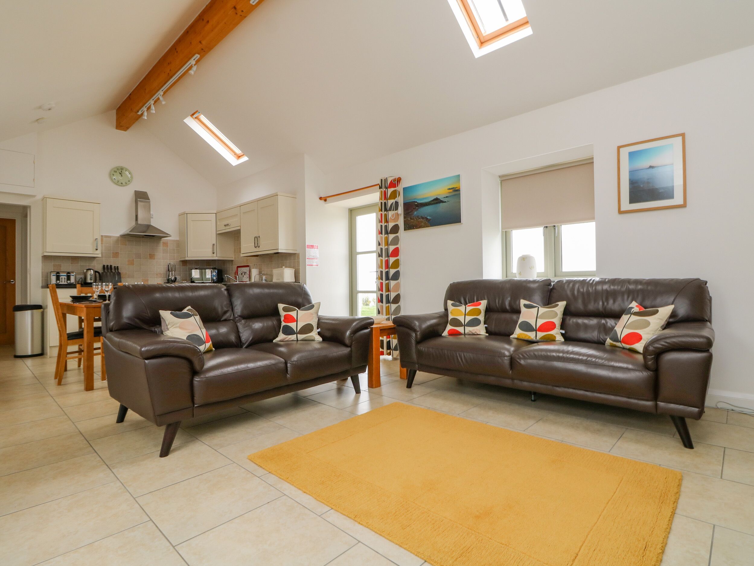 A spacious living room with two brown leather sofas, a yellow rug, and a kitchen in the background.