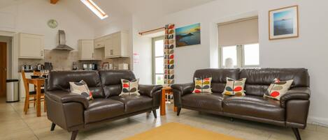 A spacious living room with two brown leather sofas, a yellow rug, and a kitchen in the background.