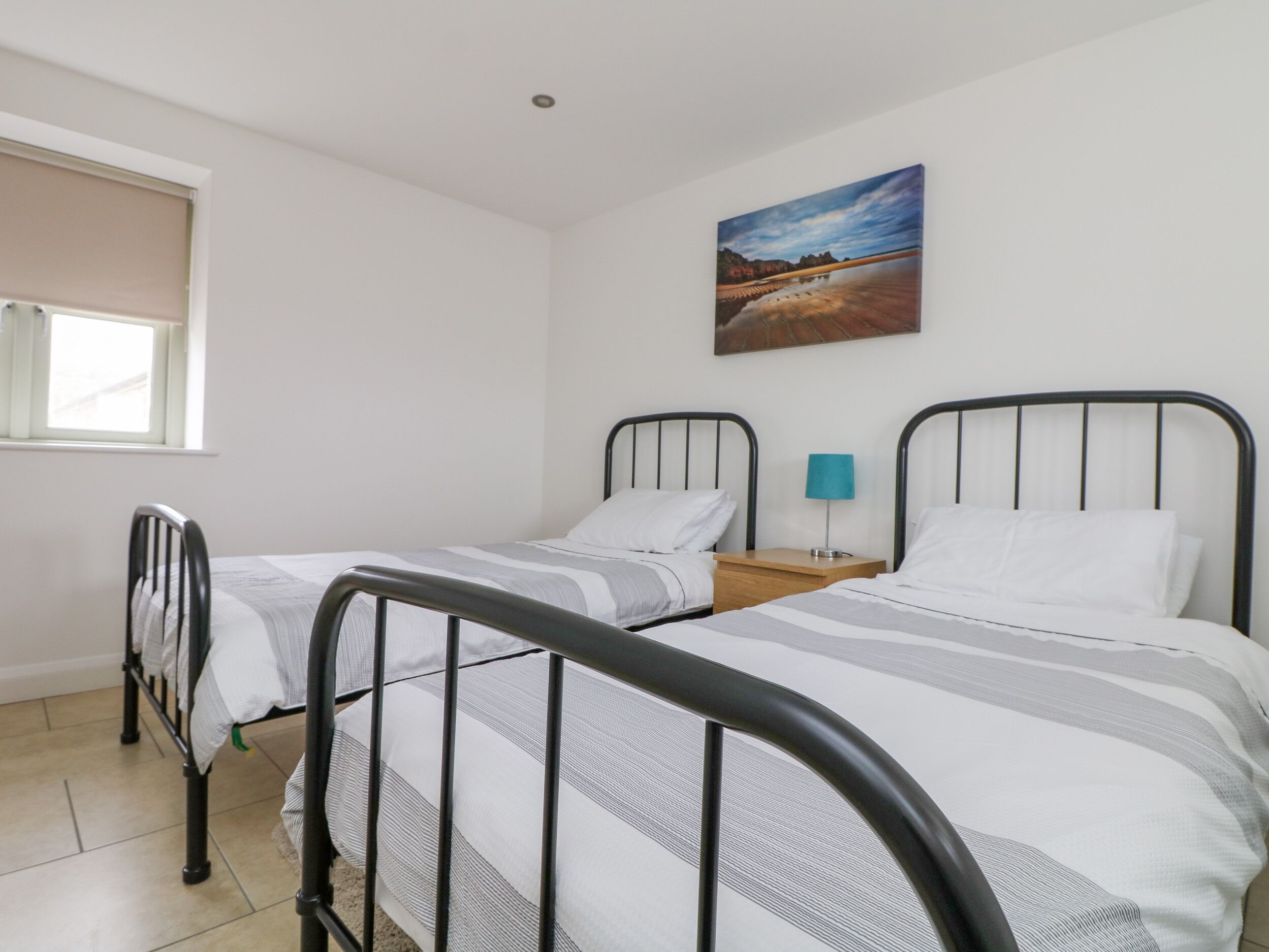 Un dormitorio con dos camas individuales, una ventana con persianas, una mesita de noche con lámpara y un cuadro en la pared.