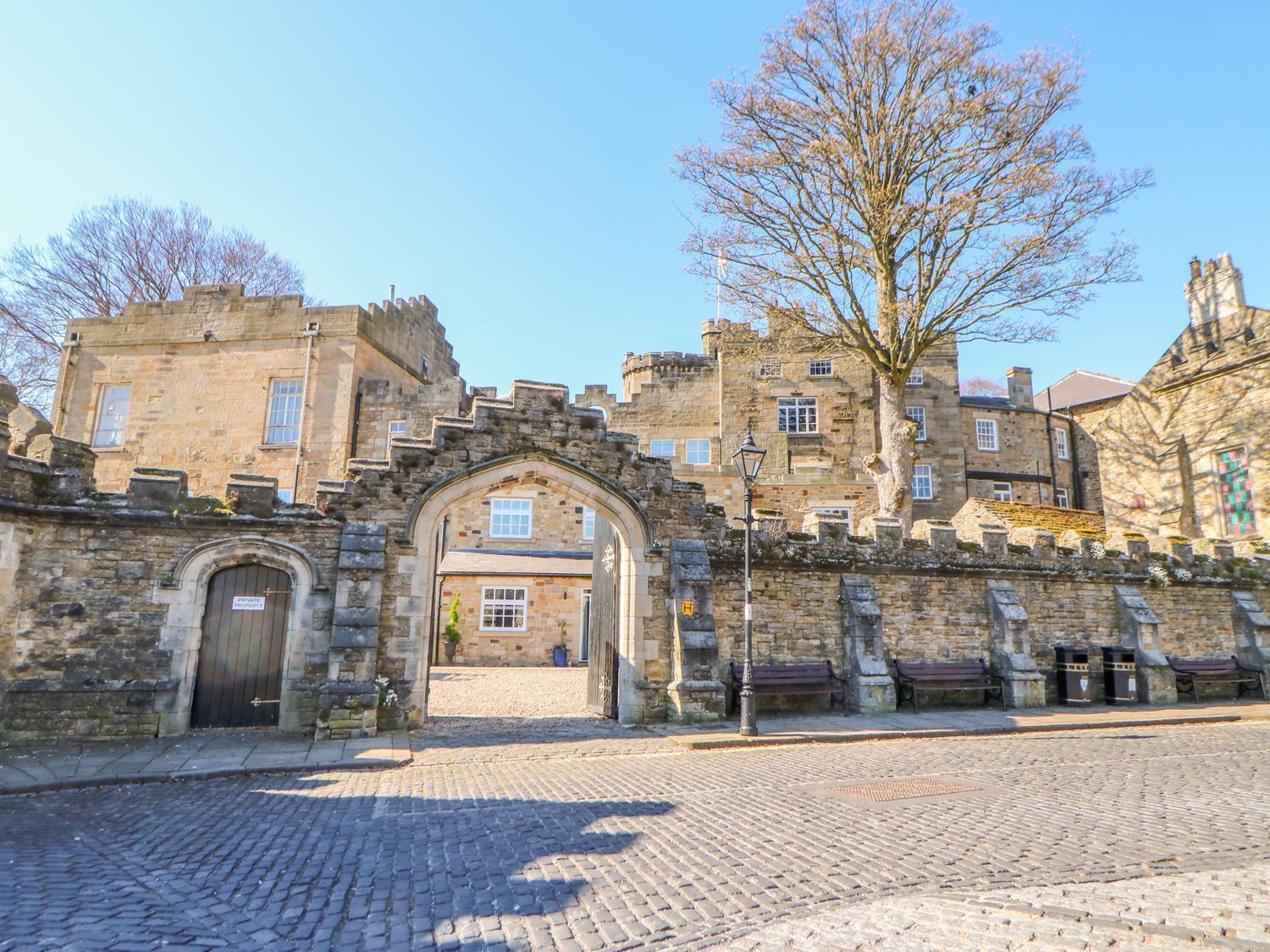 A historic stone building with a large arched entrance, a cobblestone courtyard, and a tree in the foreground.