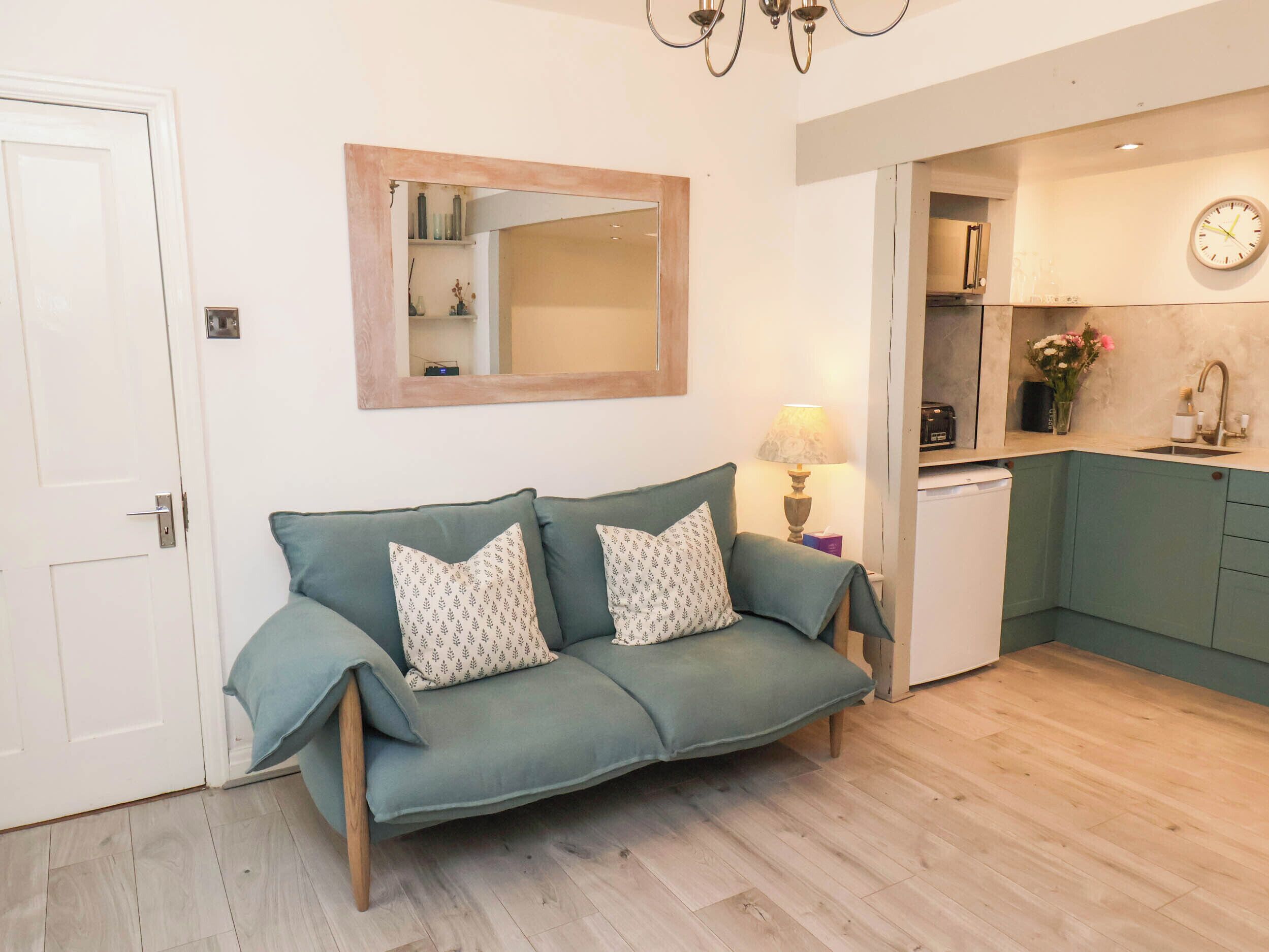 Un salon avec un canapé, un miroir, une lampe et un coin cuisine avec une horloge.