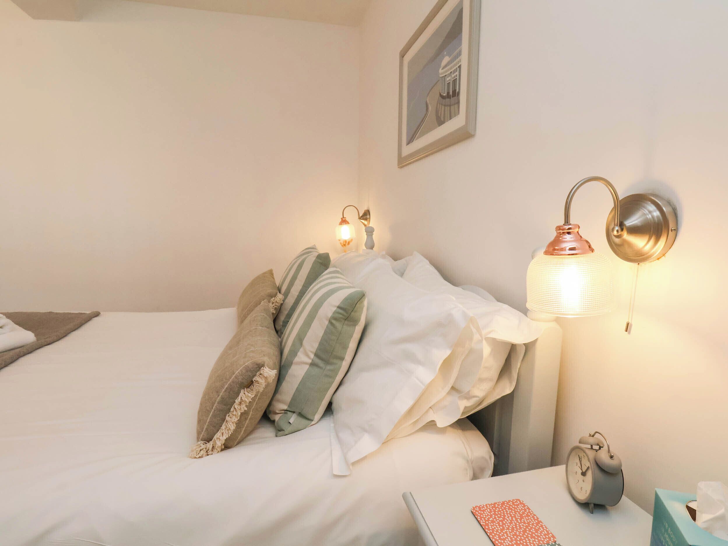 Un lit bien fait, avec des oreillers, une lampe de chevet, une horloge et un tableau encadré au mur.