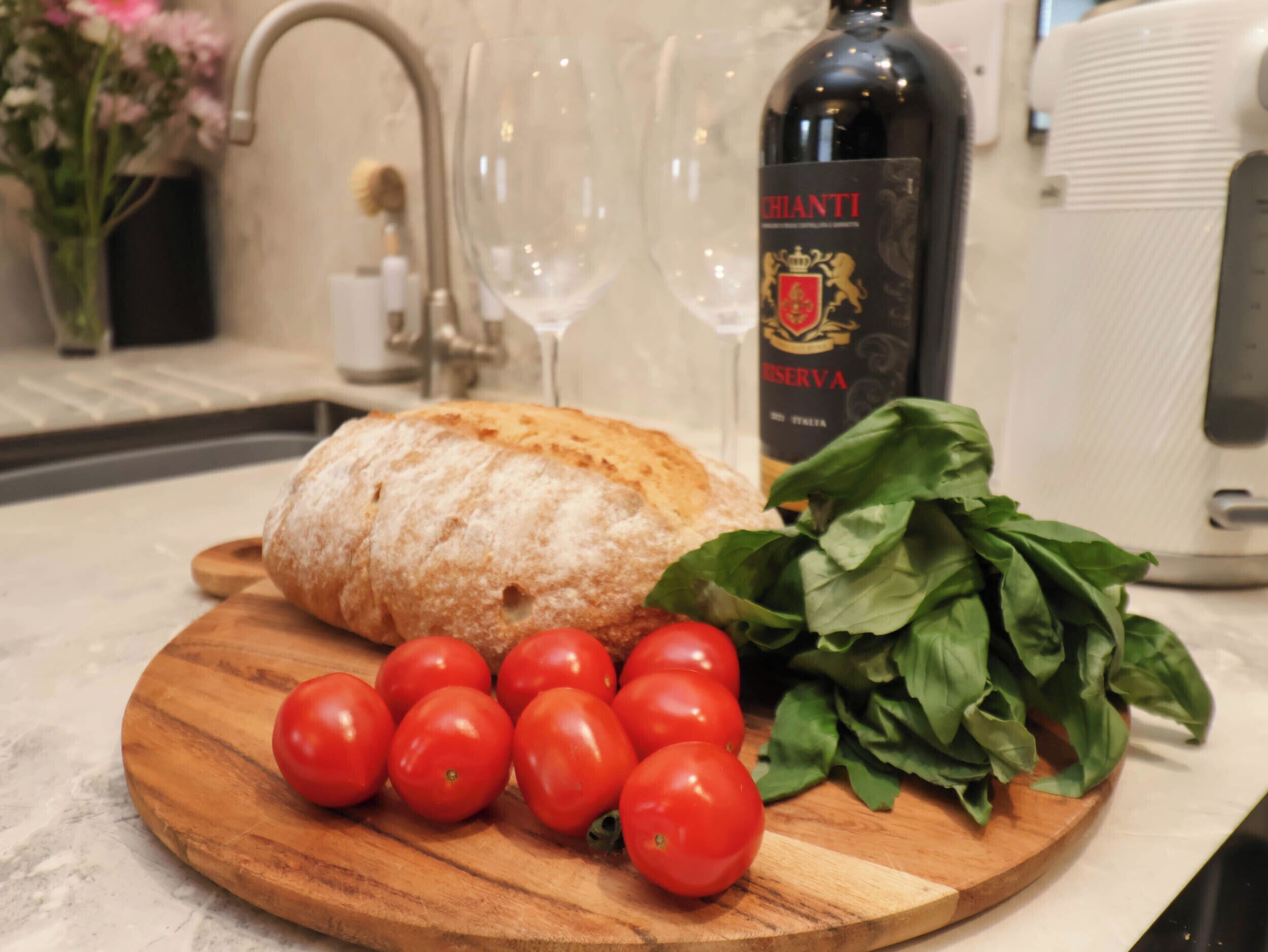 A wooden cutting board with a loaf of bread, tomatoes, and basil leaves on a kitchen counter.