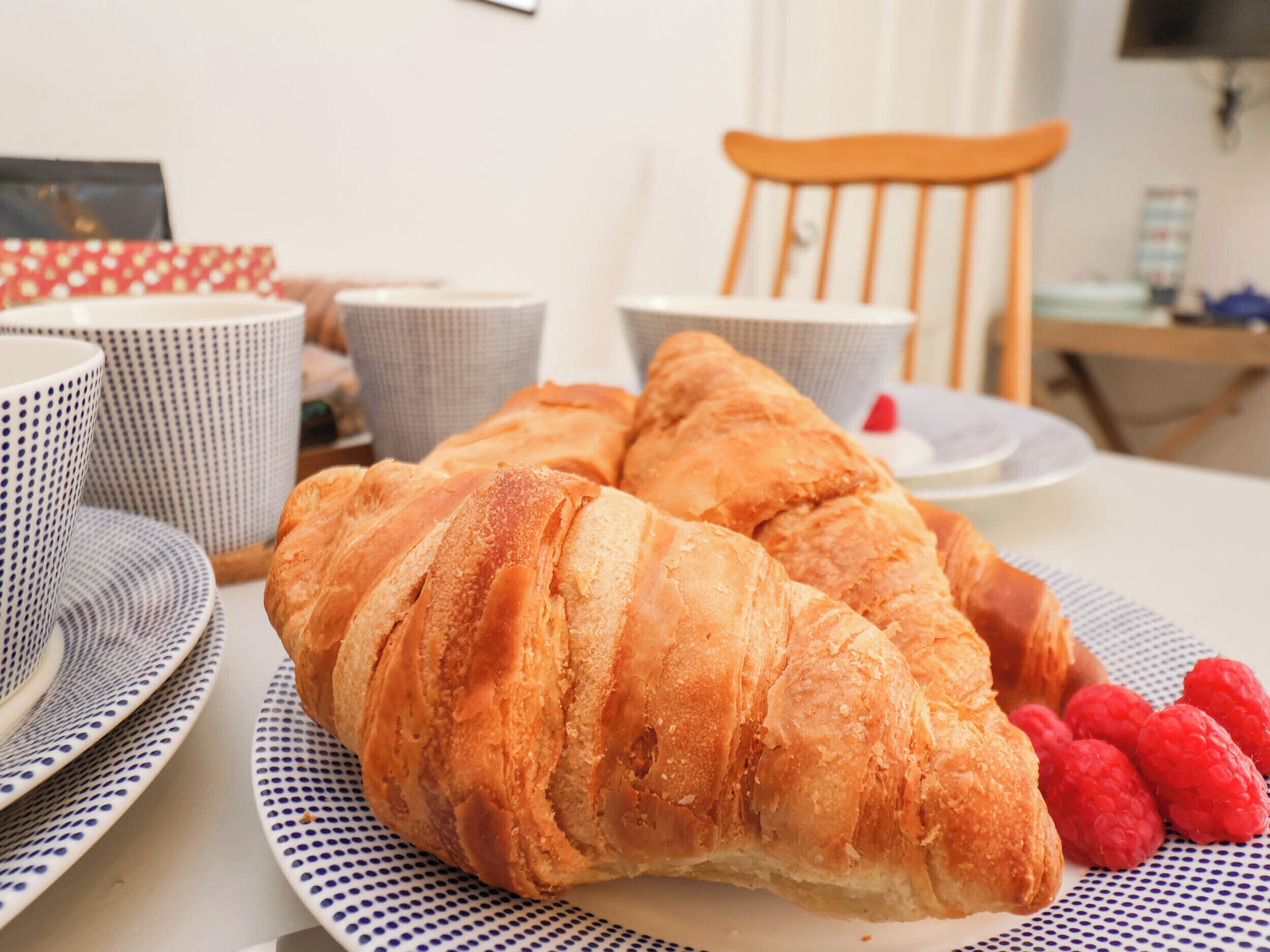 A dining table with croissants and raspberries, a wicker chair, and patterned cups.