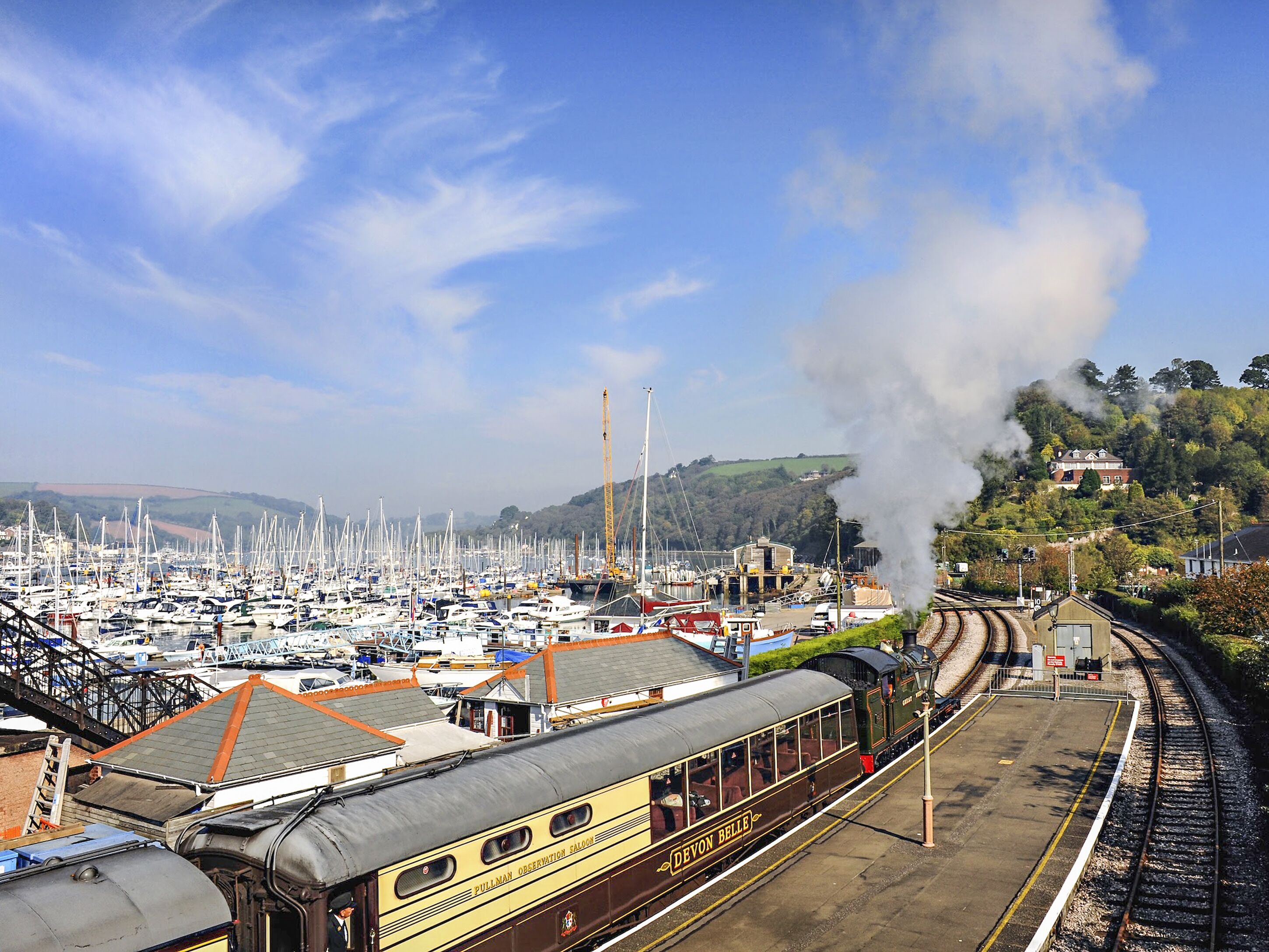 A train station with a steam locomotive emitting smoke, surrounded by a marina with numerous boats.
